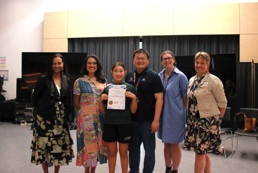 primary school student and five adults smiling to camera. The student is holding up a certificate.