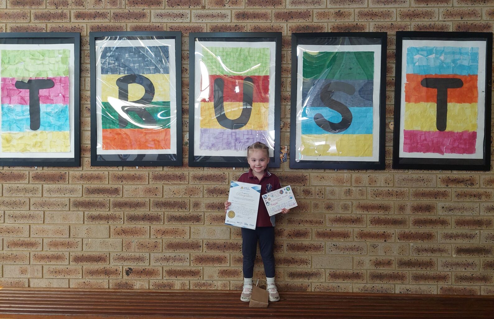 primary school student holding certificate under wall hangings that read out Trust